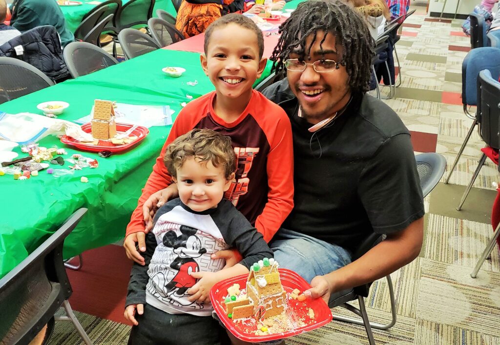 An adult and two children show off the graham cracker house they made at our 2018 gingerbread workshop event